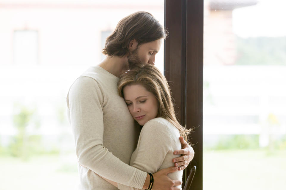 Sad young married couple embracing standing in living room opposite window at home. Sorrowful wife and husband feels unhappy, thinking about problems in relations, miscarriage, misbirth or infertility