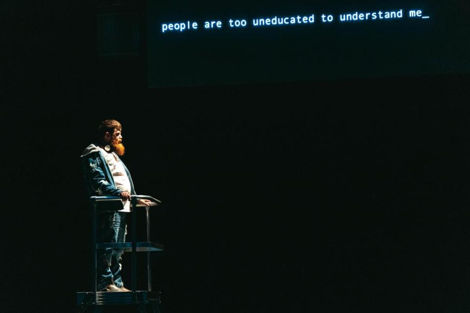 史考特在《當獵人成為獵物的影子》中，和人工智慧互動。舞台上的字幕寫著：「一般人沒有被教育如何了解我。」（背靠背劇團提供）