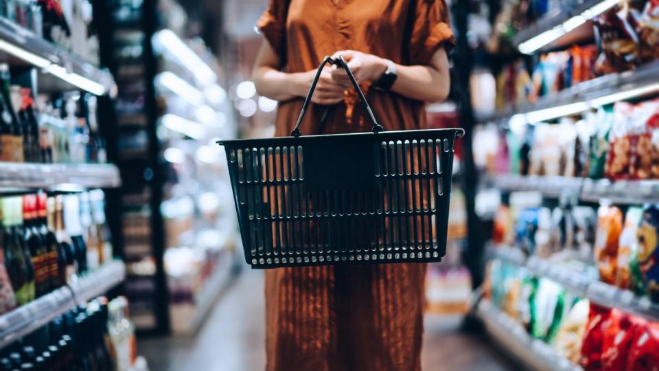 Woman in supermarket aisle holding empty grocery basket. 