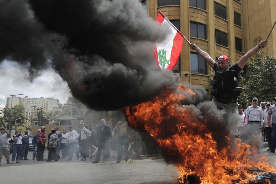 A Lebanese retired soldier holds a Lebanese national flag as he jump over burning tires during a protest in front of the government building during a cabinet meeting to discuss an austerity budget, in Beirut, Lebanon, Friday, May 10, 2019. Dozens of Lebanese military and security veterans burned tires and shouted angrily outside government offices on Friday, their second protest in less than two weeks amid fears a proposed austerity budget may affect their pensions and benefits. (AP Photo/Hassan Ammar)