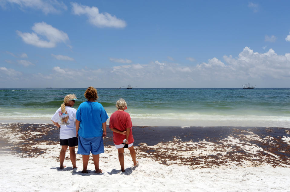 FILE- In this June 23, 2010, file photo, from left, Gina Durell, Island Durell and Linda Harrison of Pensacola, Fla., watch as crews work to clean up oil washed ashore at Pensacola Beach, Fla. Oil from the Deepwater Horizon oil spill off the coast of Louisiana washed up along the Florida Panhandle, hurting the state's tourism season that summer. (AP Photo/ Michael Spooneybarger, File)