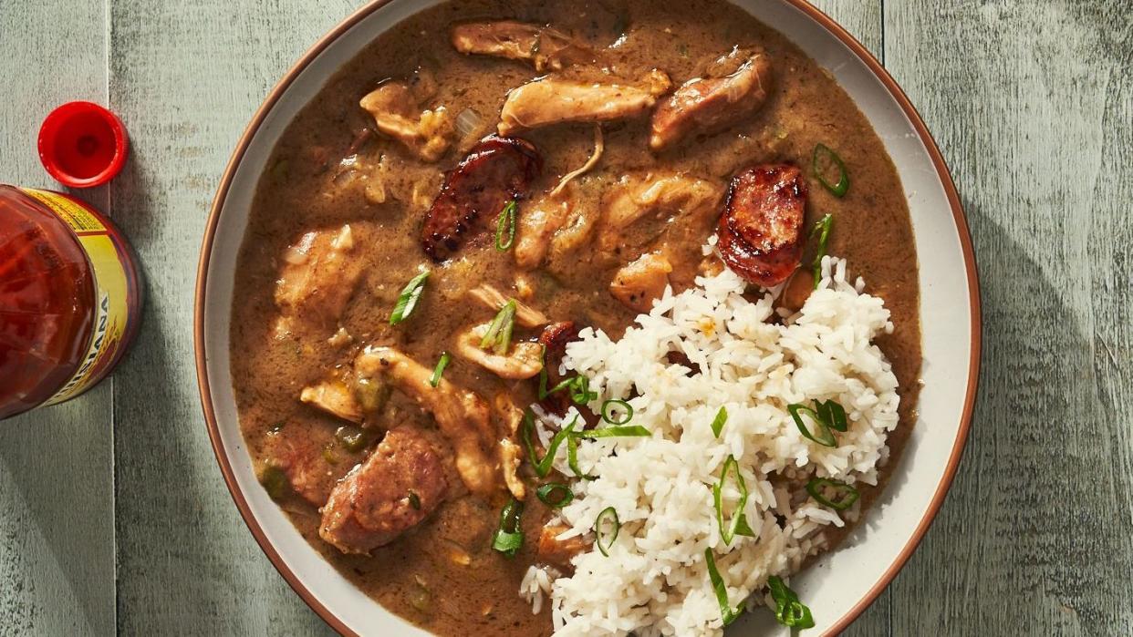 gumbo with chicken and sausage pieces in a bowl with rice topped with green onions and served with a bottle of hot sauce on the side