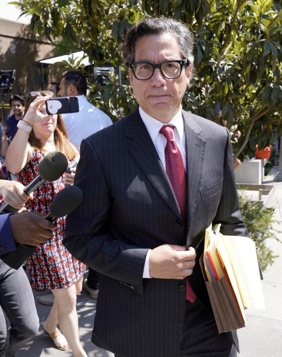 A man in a suit and tie and thick-framed glasses looks sternly at the camera