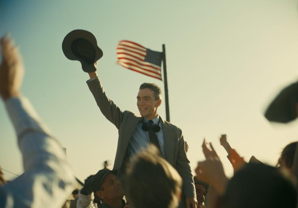 Cillian Murphy holding hat in the air as people cheer and American flag behind him