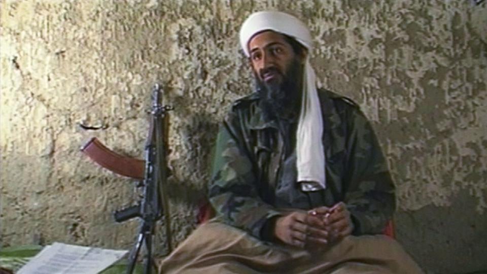 osama bin laden sits against a concrete wall and appears to be talking, he wears a white head covering, green camo jacket, and camel colored blanket on his lap, to the left rest papers and a machine gun