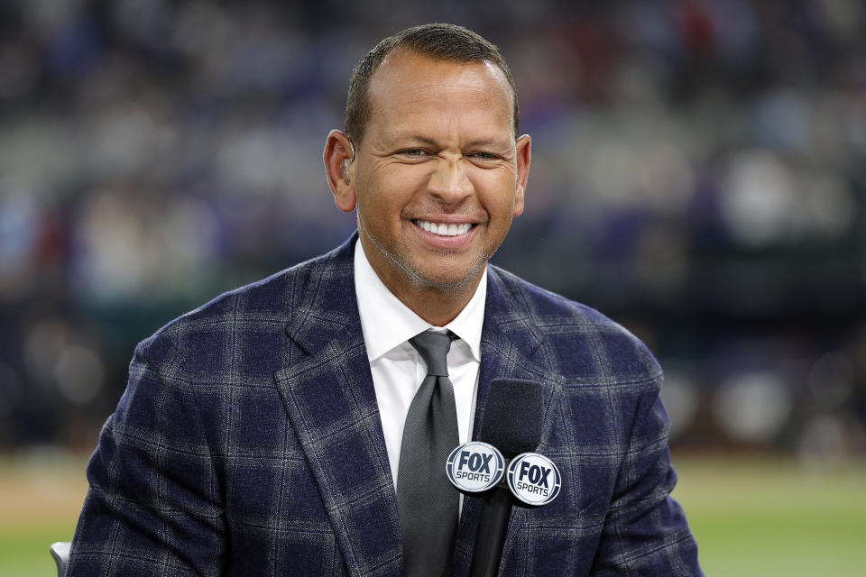 Alex Rodriguez, wearing a checked suit and tie, smiles while holding a Fox Sports microphone