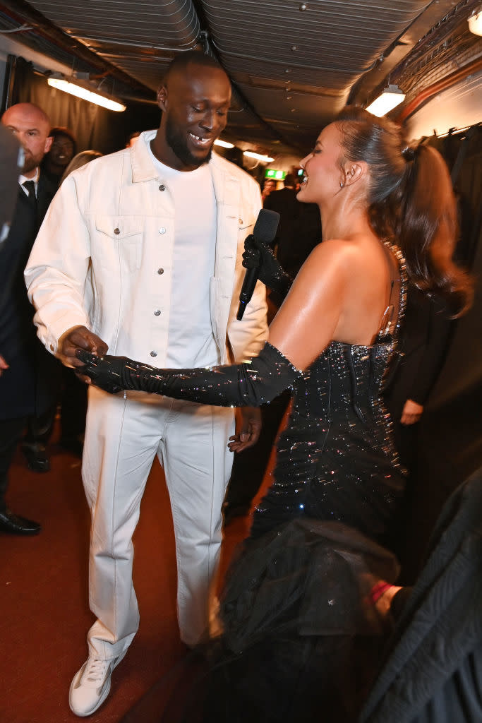 the couple interacting backstage, Stormzy in a jacket and Maya in a beaded gown