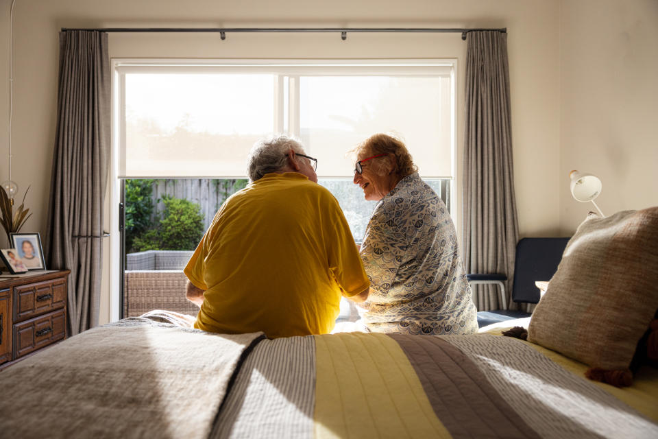 Two elderly individuals sitting on a bed facing a window, appearing to be in a conversation