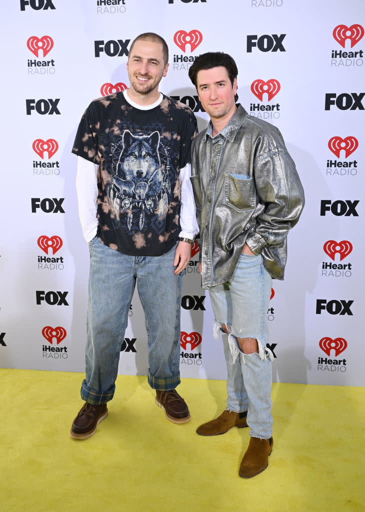 Two men posing, one in a wolf print T-shirt and jeans, the other in a metallic jacket and ripped jeans
