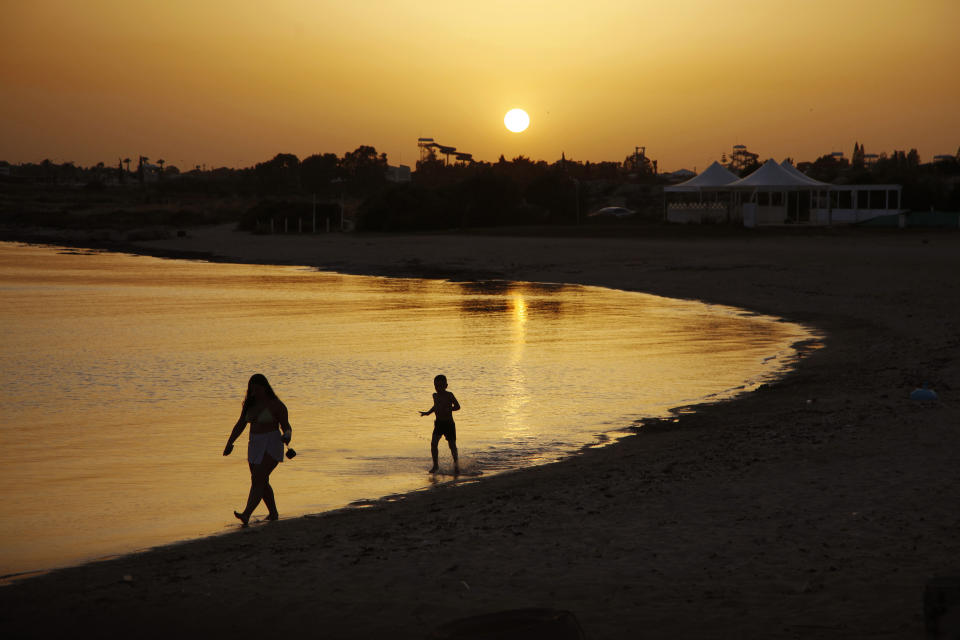 FILE - In this file photo dated Sunday, May 17, 2020, beachgoers walk in the sea water during sunset at an empty stretch of Dome beach at Makrinissos in Cyprus' seaside resort of Ayia Napa, a favorite among tourists. Cyprus' government Wednesday May 27, 2020, is pledging to cover all costs for anyone testing positive for the coronavirus while on vacation in the east Mediterranean island nation, covering the costs of lodging, food, drink and medication for COVID-19 patients and their families. (AP Photo/Petros Karadjias, FILE)