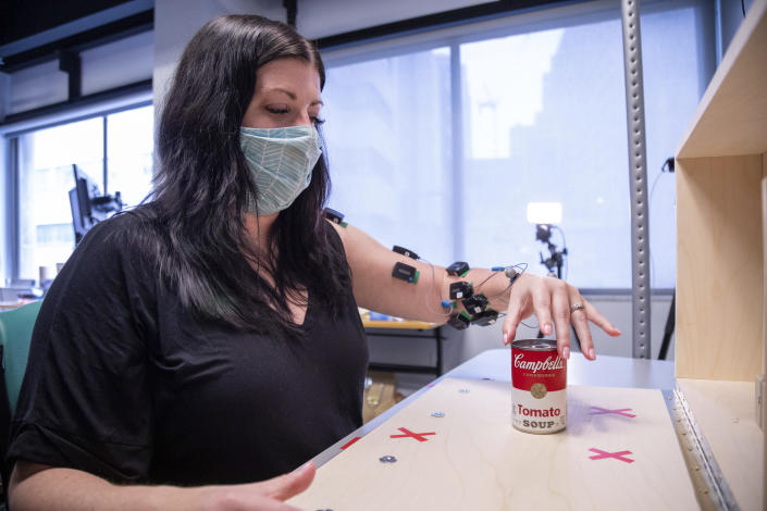 In this photo provided by UPMC and Pitt Health Sciences, research participant Heather Rendulic prepares to grasp and move a can of soup at the Rehab Neural Engineering Labs of the University of Pittsburgh on May 24, 2021. A stroke left Rendulic with little use of her left hand and arm, so she volunteered for a first-of-its-kind experiment that stimulates her spinal cord in spots that control upper limb motion. (Tim Betler/UPMC and Pitt Health Sciences via AP)