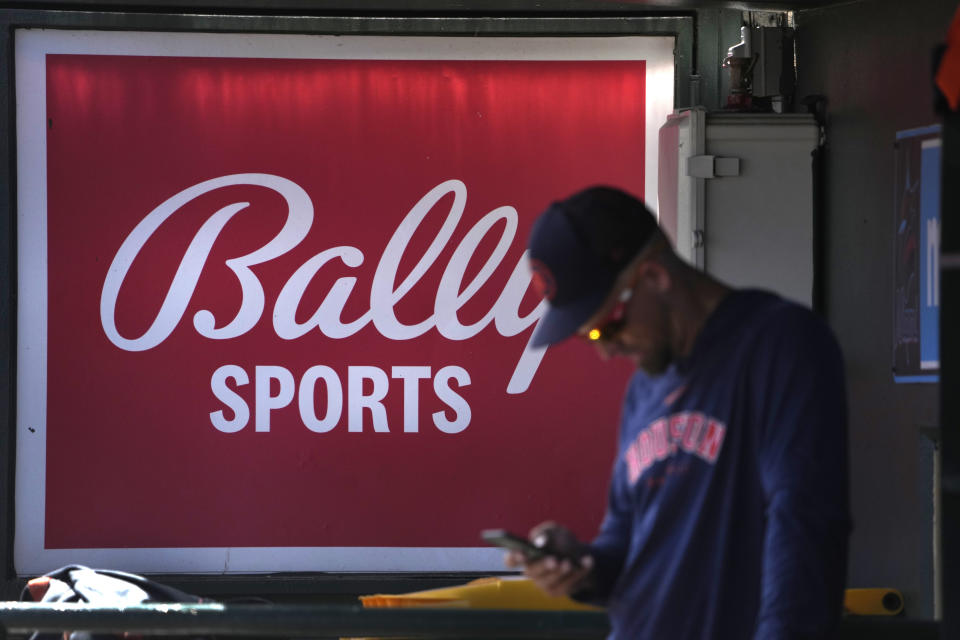 FILE - A member of the Houston Astros stands in the dugout in front of a Bally Sports sign before the team's spring training baseball game against the St. Louis Cardinals on March 2, 2023, in Jupiter, Fla. Diamond Sports Group, the largest owner of regional sports networks, filed for Chapter 11 bankruptcy protection Tuesday, March 14. The move came after it missed a $140 million interest payment last month. Diamond owns 19 networks under the Bally Sports banner. Those networks have the rights to 42 professional teams — 14 baseball, 16 NBA and 12 NHL. (AP Photo/Jeff Roberson, File)