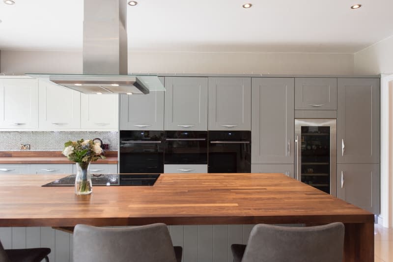 A general interior view of a large grey fitted kitchen with wooden breakfast bar island, grey velvet bar stools, black oven and microwave, induction hob, stainless steel extractor fan hood, wine beer cooler, chiller fridge, tiled floor.