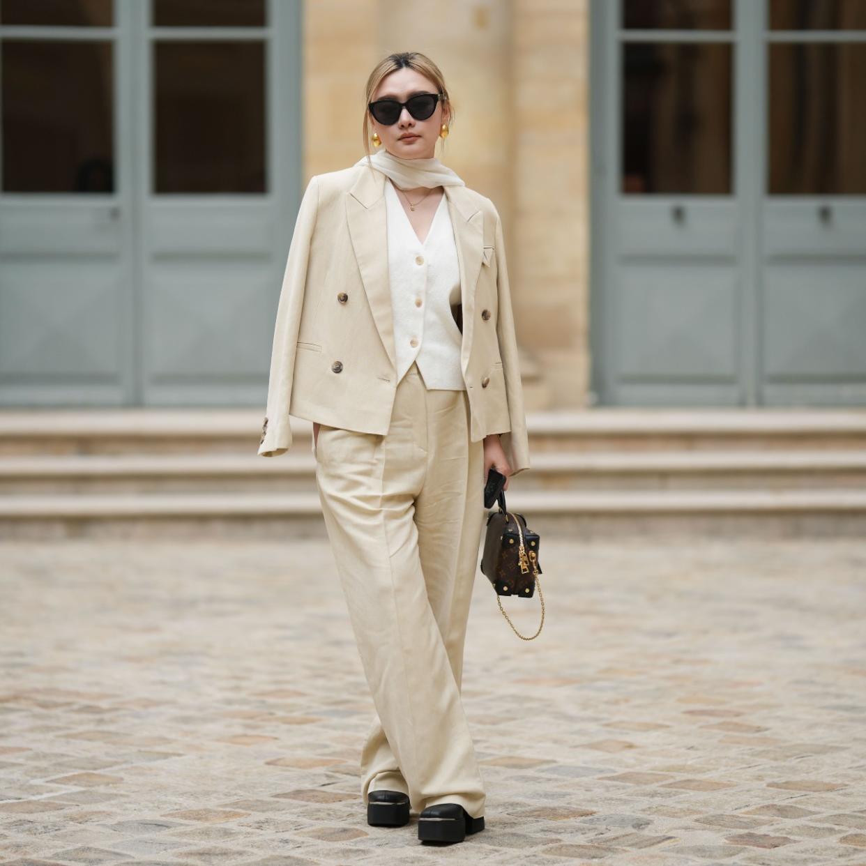  A guest wears black sunglasses, gold earrings, a white silk scarf, a gold chain pendant necklace, a white tweed buttoned V-neck gilet jacket, a beige linen buttoned blazer jacket, matching beige large suit pants, a brown shiny LV monogram print pattern in coated canvas zipper Petite Malle handbag from Louis Vuitton, black shiny leather platform soles / block heels ankle boots , outside the COS show, on April 26, 2023 in Paris, France. 