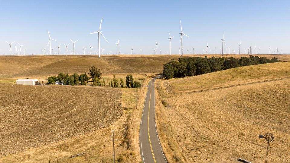 A long stretch of road in California farmland leads into the horizon, which is dotted with wind turbines.