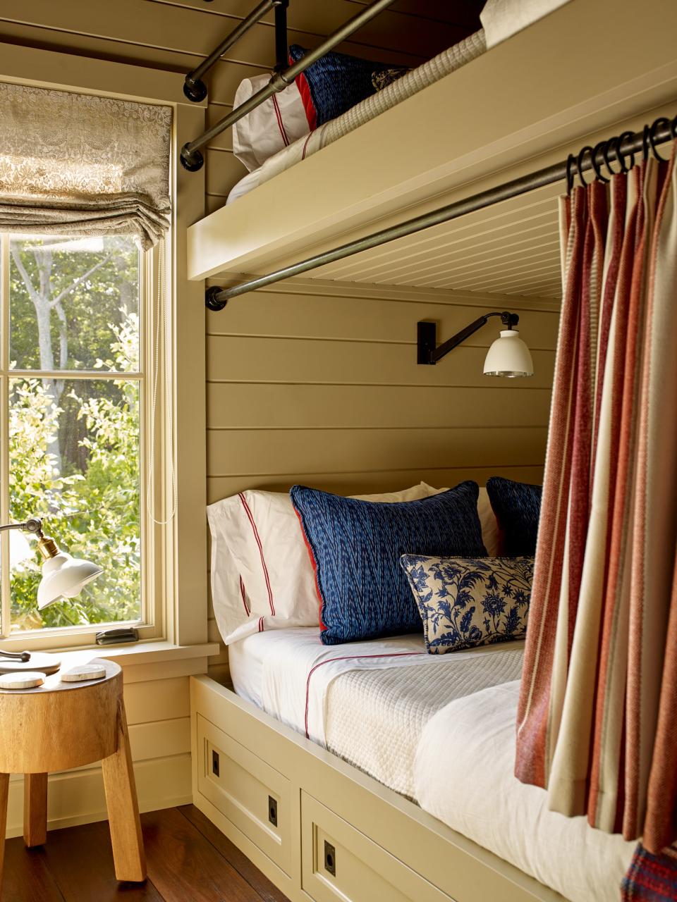 Inspired by the tight living quarters seen on boats near the house, Lippmann designed built-in drawers that hold extra blankets in this bunk room. Beds with iron pipe privacy rods by Cumberland Ironworks and custom curtains in wool by Duralee can be seen. The paint color is Light Stone by Farrow & Ball.