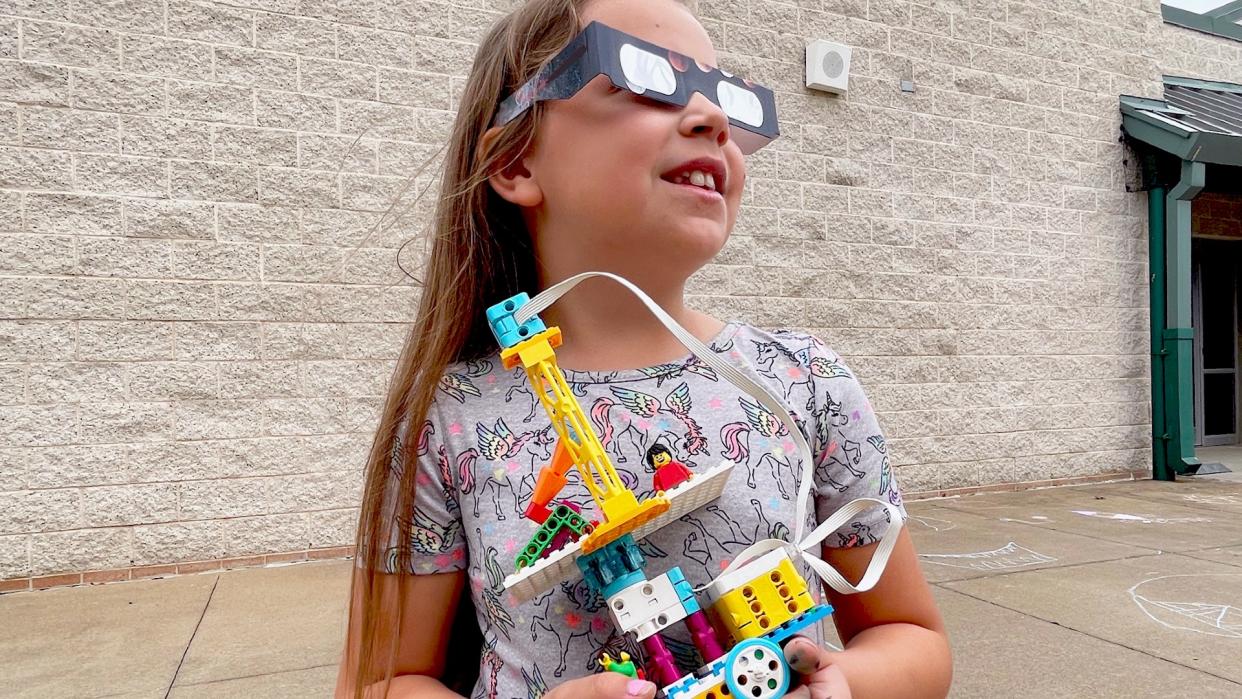  A young girl wears eclipse glasses while looking up and holding a lego creation. 