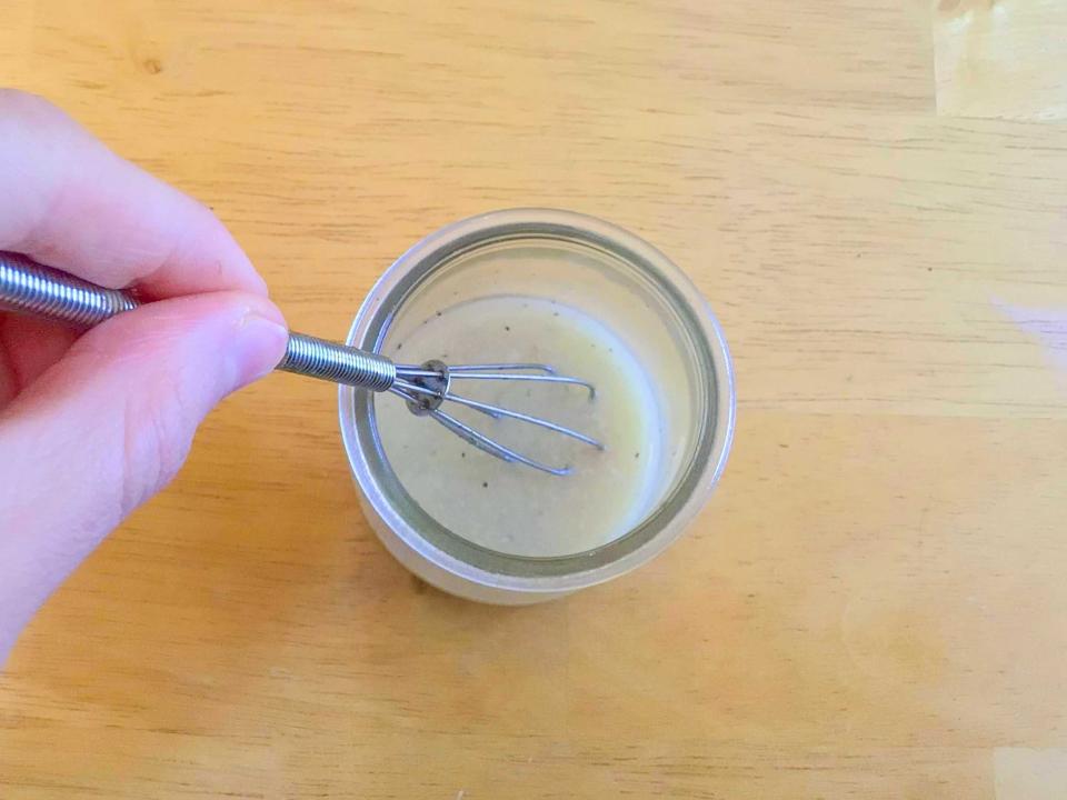 A hand holding a small whisk stirring a vinaigrette on a wooden table. The vinaigrette is a pale beige color.