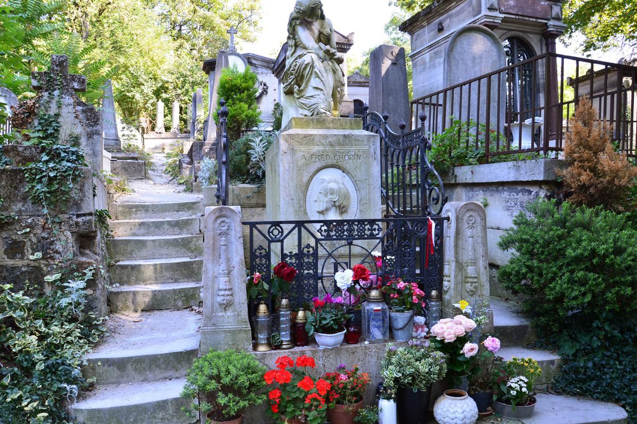 Frederic Chopin's grave in the Père Lachaise Cemetery, Paris with steps on both sides and fenced off with several candles with light shining through the background, several other gravesites