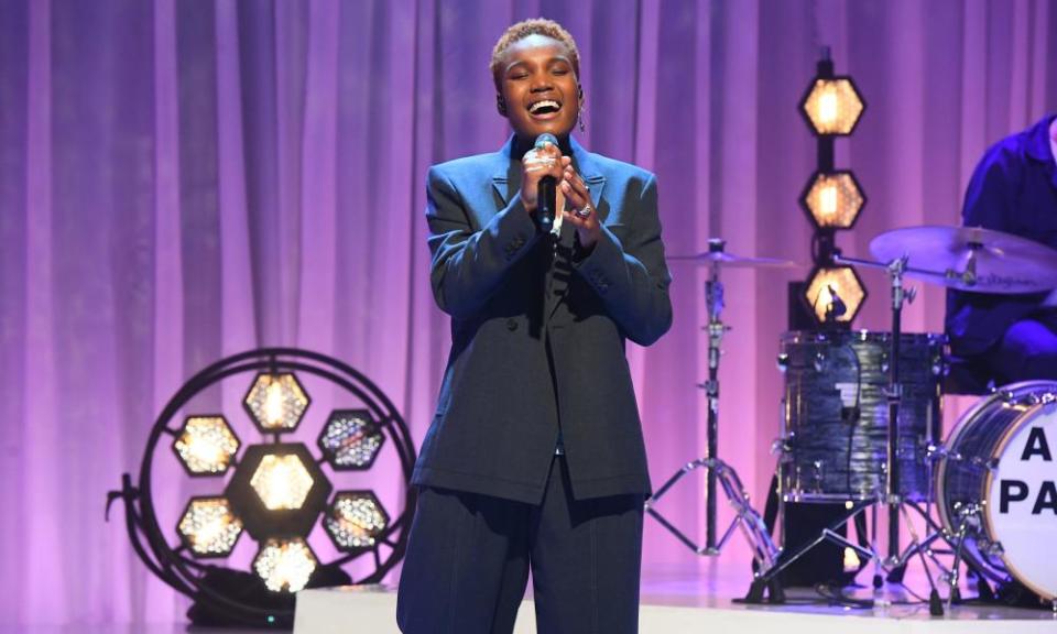 Arlo Parks onstage in a blue suit, singing, with two arrays of floor mounted spotlights behind her and her drummer partly visible to the right