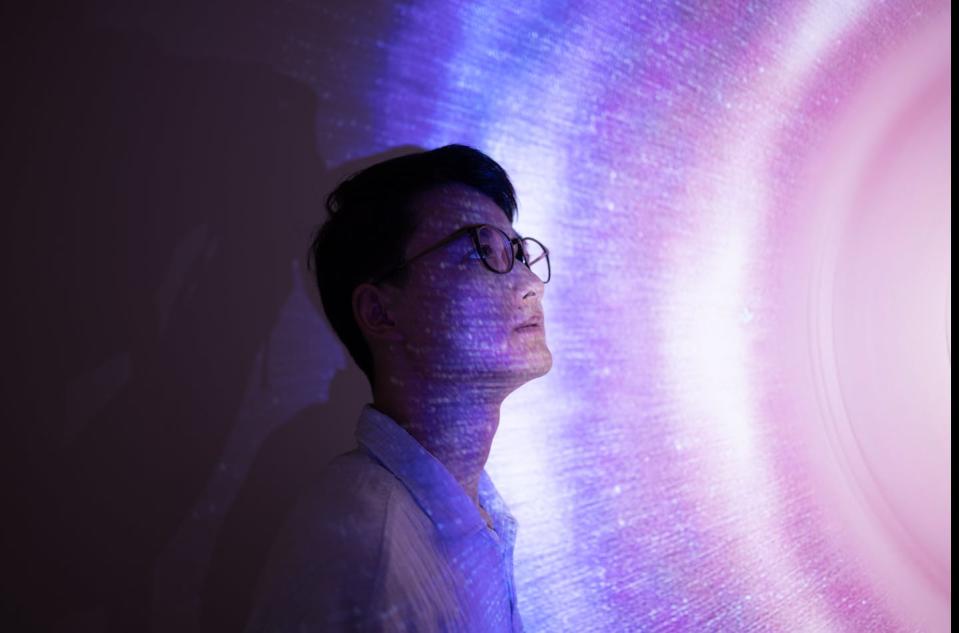 An Asian man wearing glasses stares seriously into space, standing against a holographic background in pink and blue tones.