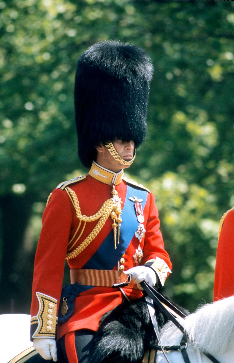 King Charles III wearing a red military uniform and a royal guard hat while riding a horse.