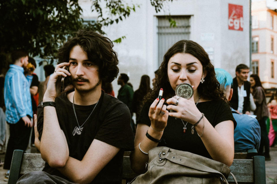 Two individuals sitting on a bench; the person on the left is on the phone, and the person on the right is applying lipstick using a handheld mirror