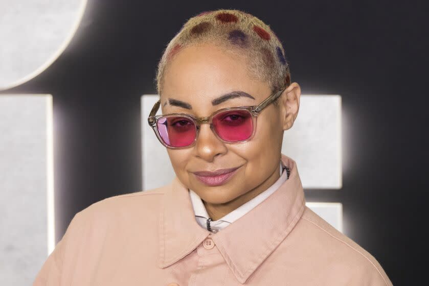 Raven-Symoné with a patterned buzzcut wearing pink sunglasses and a pink jacket smiling in front of a black background