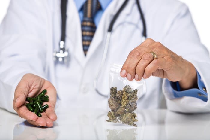 A physician with a stethoscope around his neck and holding a baggie of dried cannabis in his left hand and cannabis oil capsules in his right hand.