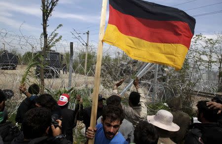 A migrant carries a German flag in front of Macedonian police as they stand guard behind a fence near a makeshift camp at the Greek-Macedonian border near the village of Idomeni, Greece, April 11, 2016. Reuters/Alexandros Avramidis