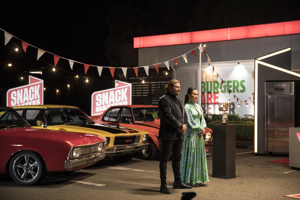 Snackmasters hosts Poh Ling Yeow and Scott Pickett stand in front of hot rod cars at the Whooper Burger Cook-Off at the Warriewood Hungry Jacks, Sydney