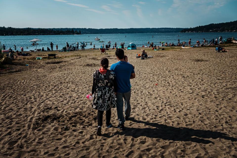 A woman and a man holding a child, seen from behind, walk on a beach toward other beachgoers