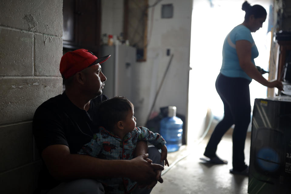 FILE - In this Jan. 28, 2020, file photo, Juan Carlos Perla sits with his son, Joshua Perla, as his wife, Ruth Aracely Monroy, right, stands in their home on the outskirts of Tijuana, Mexico. After fleeing violence in El Salvador, the family is among 60,000 U.S. asylum seekers returned to Mexico to wait while their claim makes its way through the U.S. court system. The Trump administration has suspended immigration court hearings for asylum-seekers waiting in Mexico through June 1, bowing to public health concerns arising from the coronavirus while extending a state of limbo those locked down in Mexican migrant shelters. (AP Photo/Gregory Bull, File)