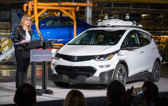 Mary Barra is shown standing next to a white Chevrolet Bolt EV with visible self-driving sensor hardware at an event at GM's Orion Assembly plant in June 2017.