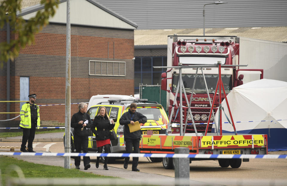 People walk past a police officer at the scene after a truck, seen in rear, was found to contain a large number of dead bodies, in Thurrock, South England, early Wednesday Oct. 23, 2019. Police in southeastern England said that 39 people were found dead Wednesday inside the truck container believed to have come from Bulgaria. (Stefan Rousseau/PA via AP)