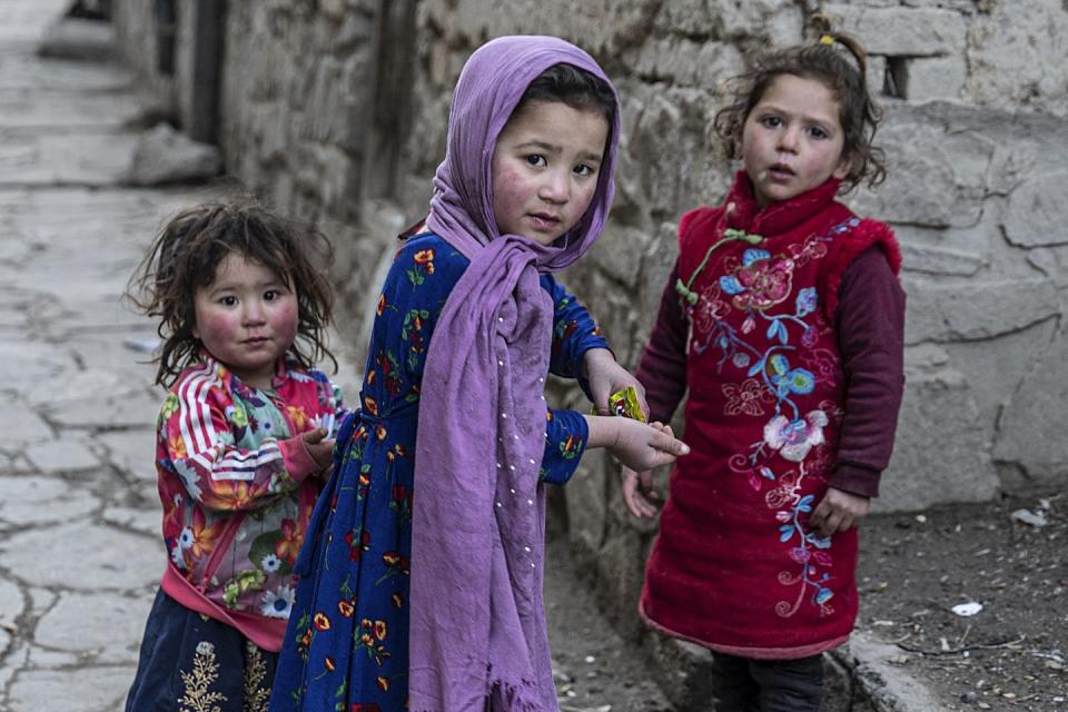 Three girls in colorful garments eat snacks on a street.