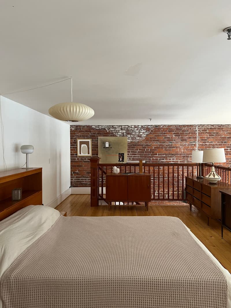 Wood furniture and bed with tan textured blanket in bedroom with exposed brick wall.