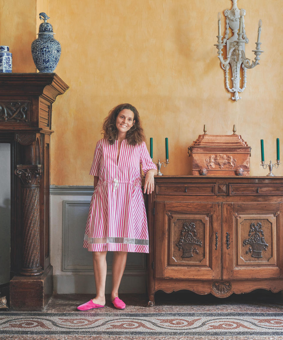 French house with authentic  yellow walls, large antique sideboard and fire place, mosaic tiled floor, pottery, candlesticks, wall light, owner stood in pink stripe dress, pink shoes