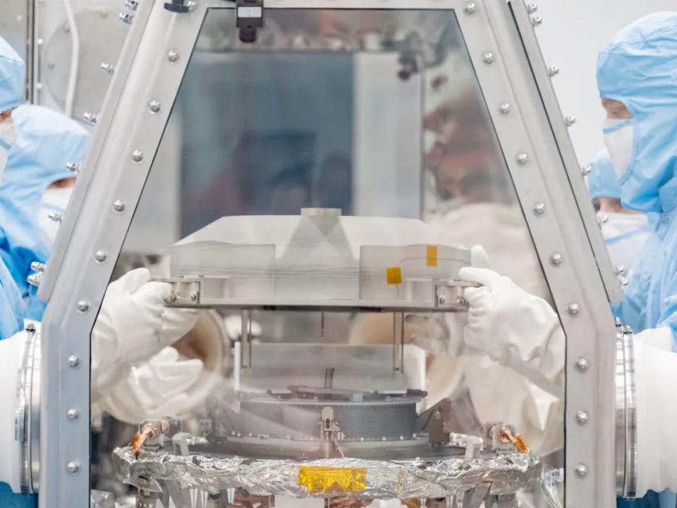 Several people in blue protective suits and mask use white gloves in a clean room containing a sample of the Bennu asteroid from NASA's OSIRIS-REx mission