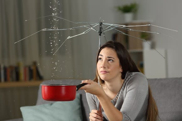 Young woman under an umbrella that has been stripped of its canopy, collecting rainwater in a cooking pot with a wistful eye on the ceiling.