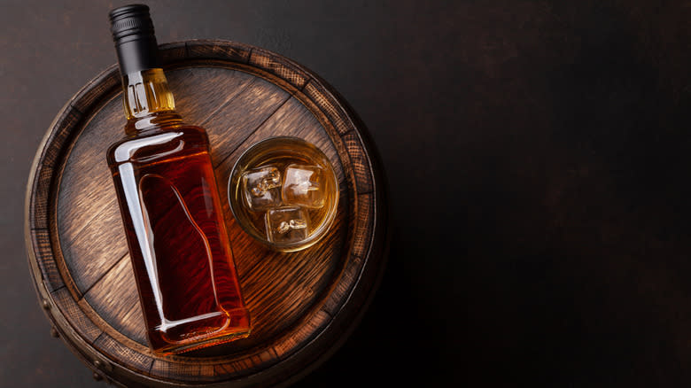 bottle of bourbon next to glass with ice on a barrel