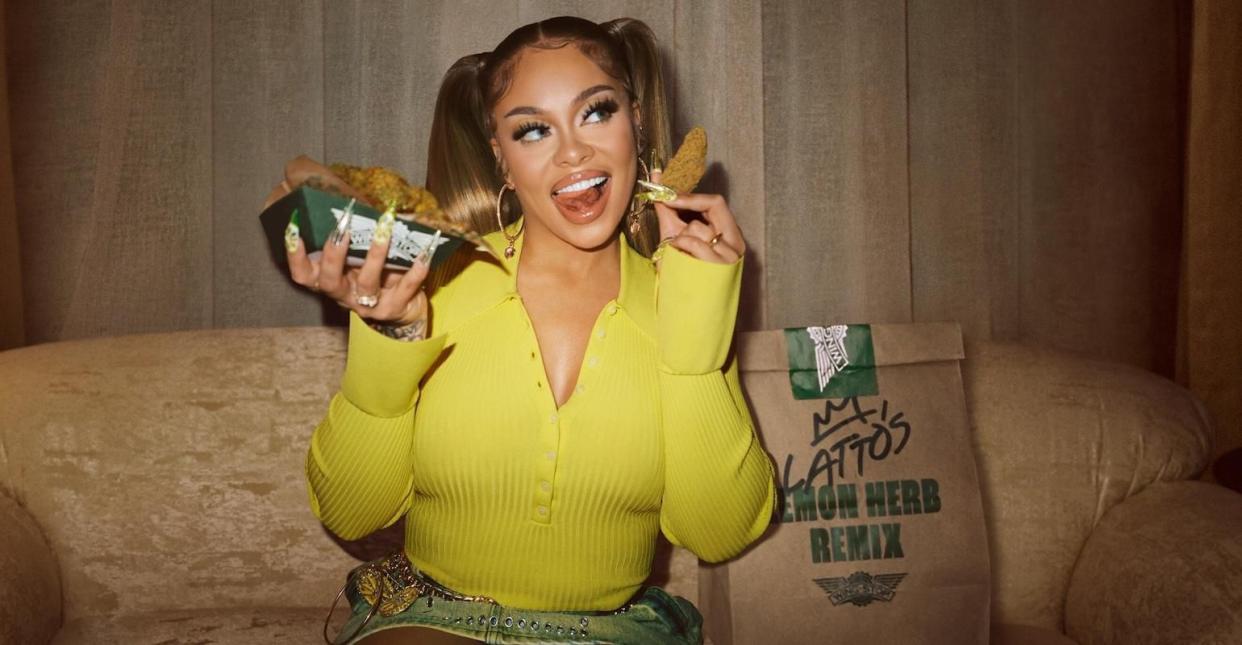 The American rapper Latto with a Wingstop bag with the words "Latto's Lemon Herb Remix" on it. She's wearing a bright yellow sweater and holding wings in both hands.