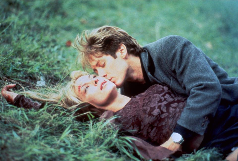 A man in a coat kisses a woman with a visible face wound on a grassy hill