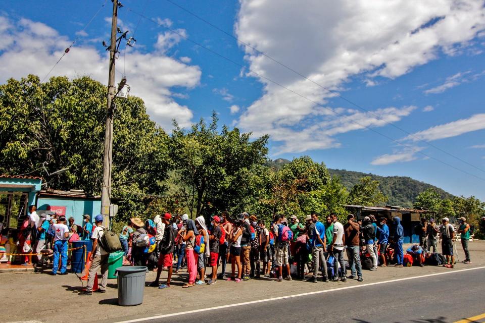 Migrants in a line wearing face masks in a verdant, warm-weather climate