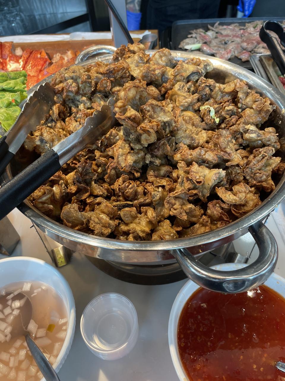 A large stainless steel pot is filled with deep-fried mushrooms, with serving tongs resting on the side. Bowls of dipping sauces and condiments are next to it