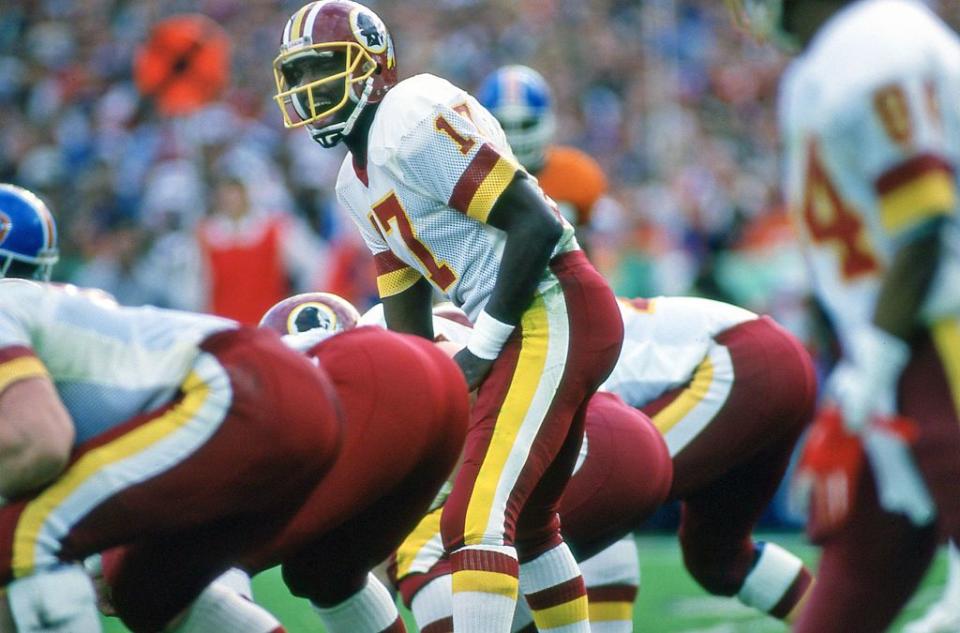 doug williams prepares to take a snap during football game, he stands in a white, red and yellow uniform with a helmet as men in matching uniforms crouch nearby