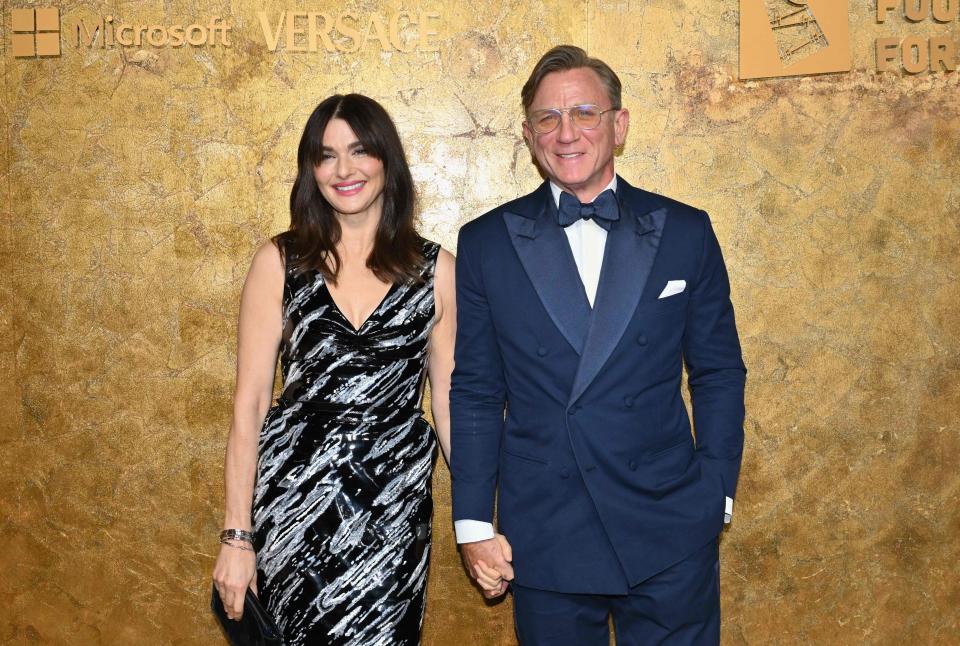 Rachel Weisz, wearing a black-and-white dress, and Daniel Craig, wearing a navy double-breasted suit with a matching bowtie, smile and hold hands on the red carpet