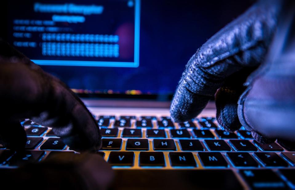 A hacker wearing black gloves who's typing on a back-lit keyboard in a dimly-lit room. 