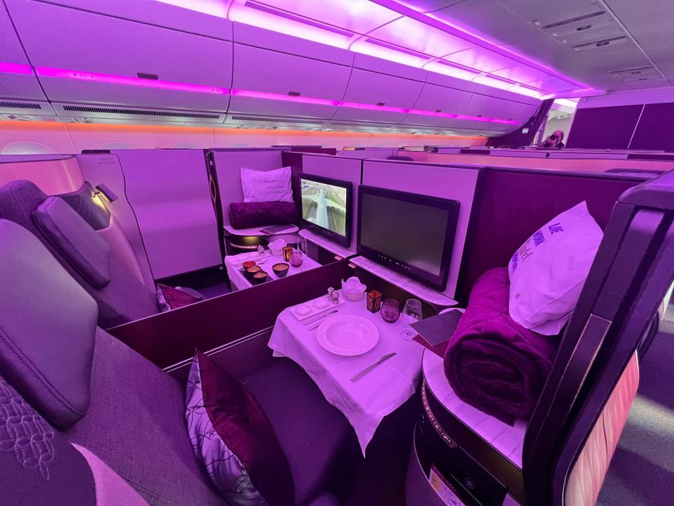 A view of two Qatar Airways business class seats side-by-side with a purple hue, and dinnerware laid out.