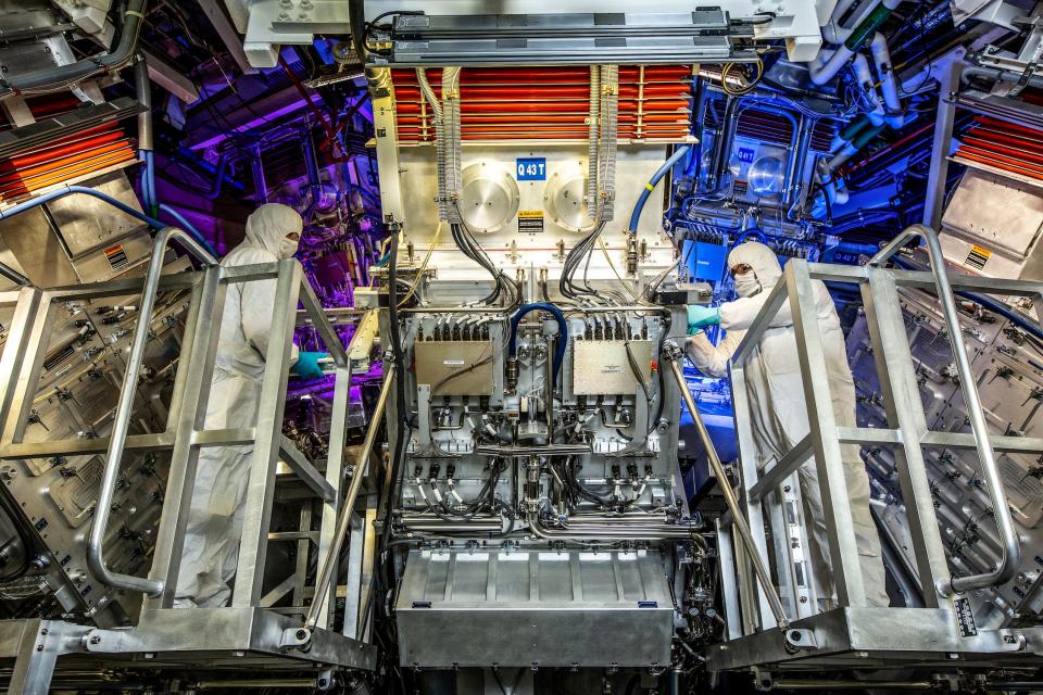 Two people in white coverall suits look at the machinery at the National Ignition Facility Target Area.